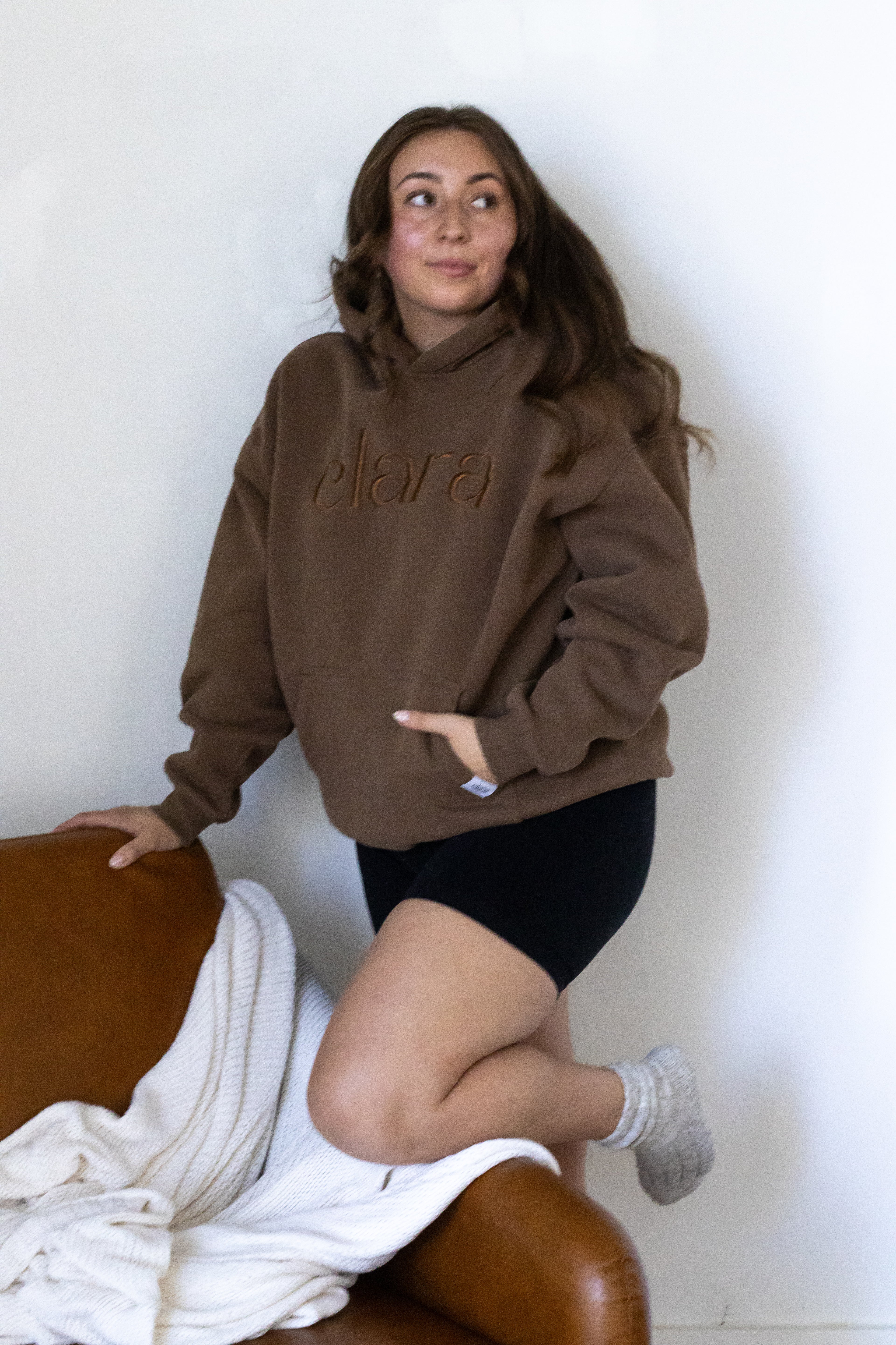 Person wearing a brown hoodie with 'Elara' text, kneeling on a chair against a white wall.