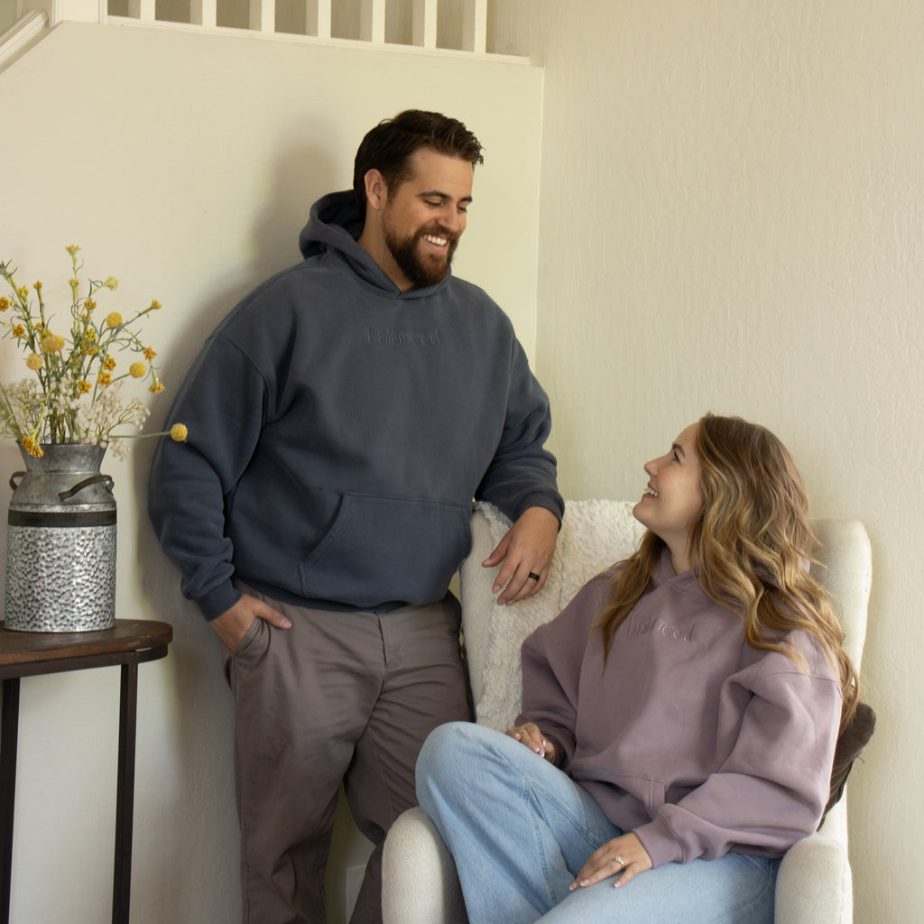 Man and woman sitting together in a cozy living room wearing hoodies with 'balanced' text