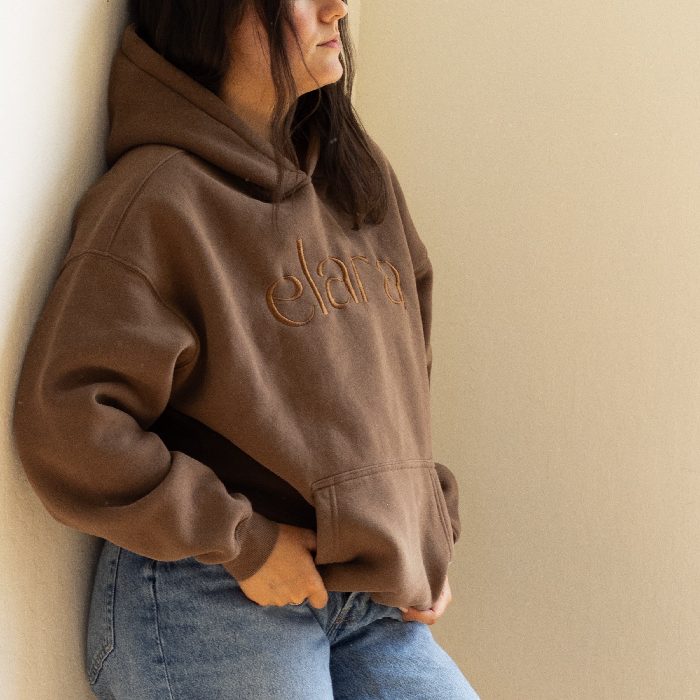 Person wearing a brown hoodie with 'elara' logo against a beige wall